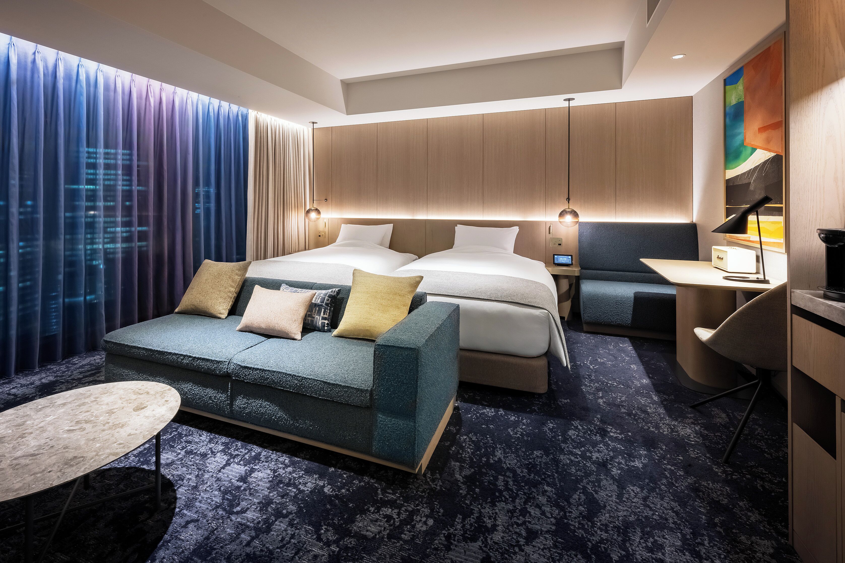 Ein modernes Hotelzimmer mit einem großen Bett, einer Sitzecke, einem Schreibtisch und Blick auf die Stadtlandschaft durch das Fenster.