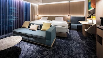 A modern hotel room with a large bed, a sofa, a desk, and a view of the cityscape through the window.