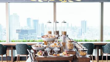 A buffet setup with various food items and containers, a city view through large windows, and modern furniture.