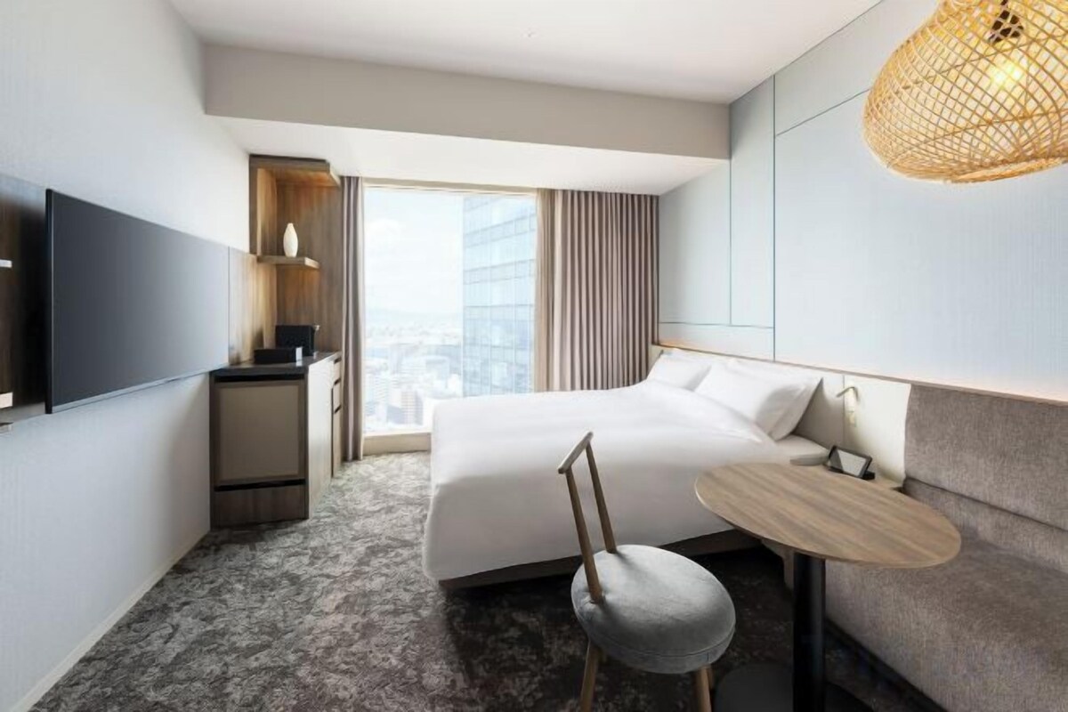 A hotel room with a large bed, a desk, a chair, a television, and a view of the city.