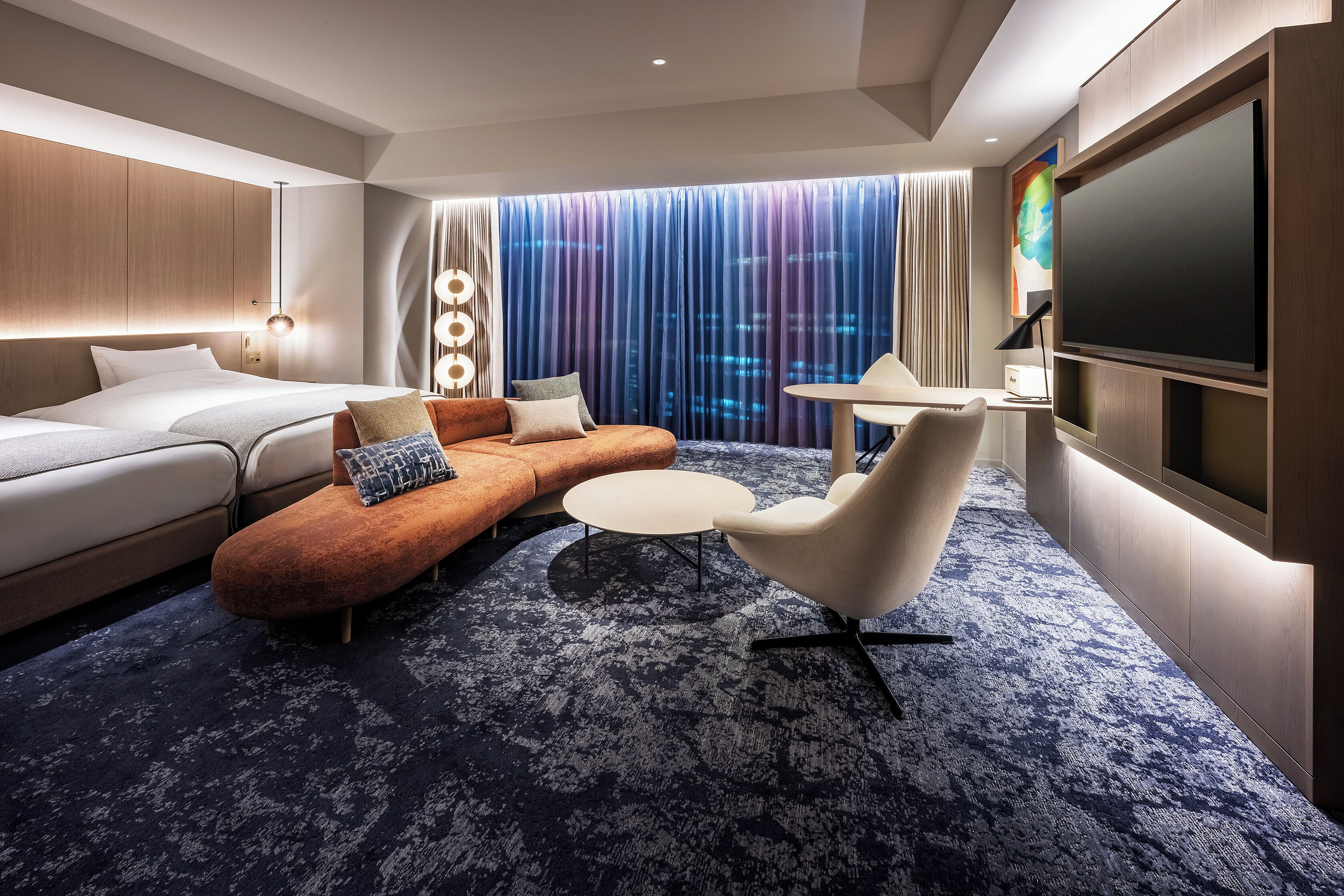 A modern hotel room with a sofa, ottoman, round coffee table, and a flat-screen TV.