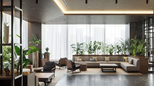 A modern living room with a sectional sofa, a coffee table, and multiple chairs. There are large windows with sheer curtains and several potted plants.