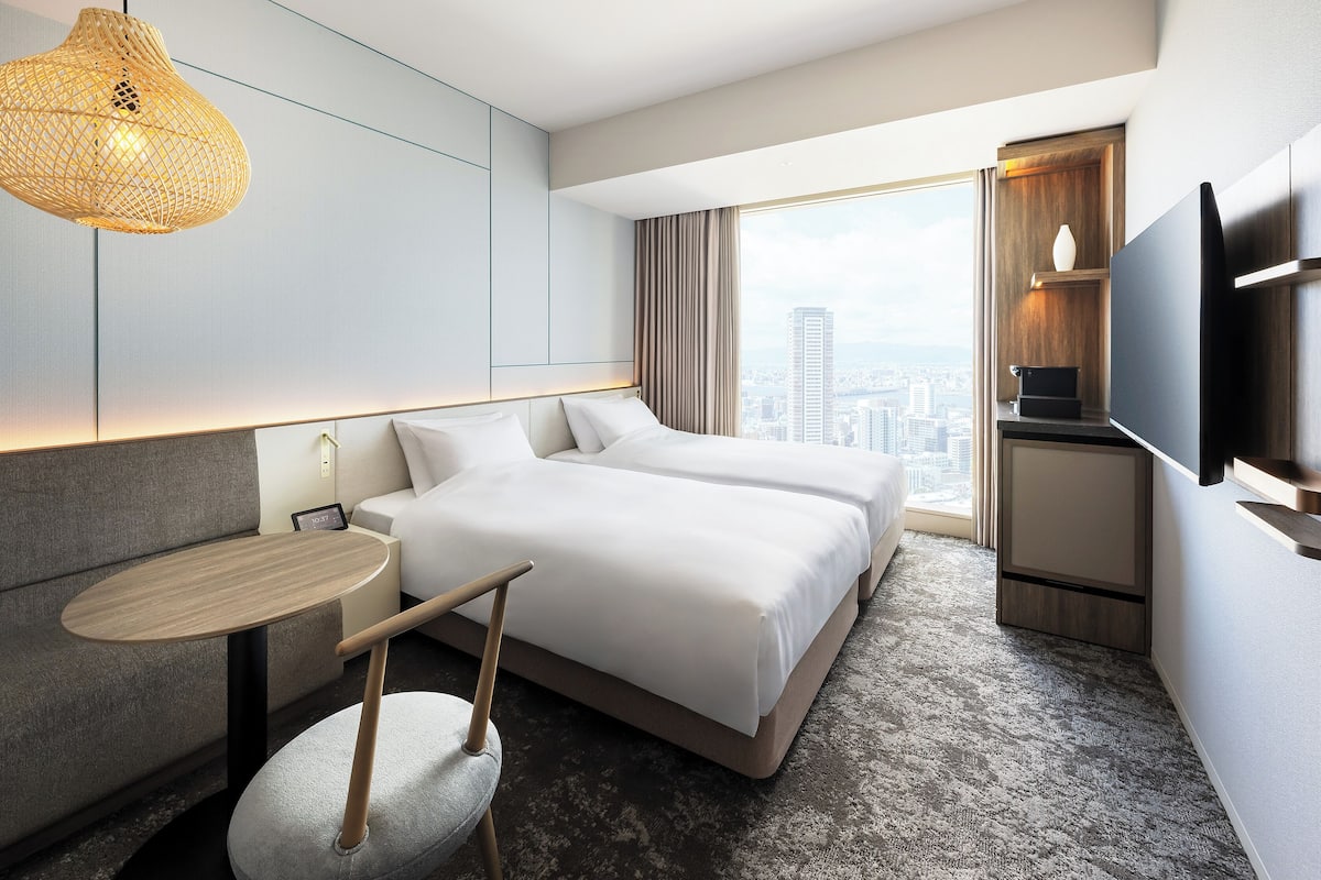 Quarto de hotel moderno com uma cama grande, uma mesinha, uma cadeira, uma televisão e vista para a cidade.