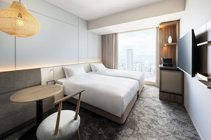 A modern hotel room with a large bed, a small table, a chair, a TV, and a city view.