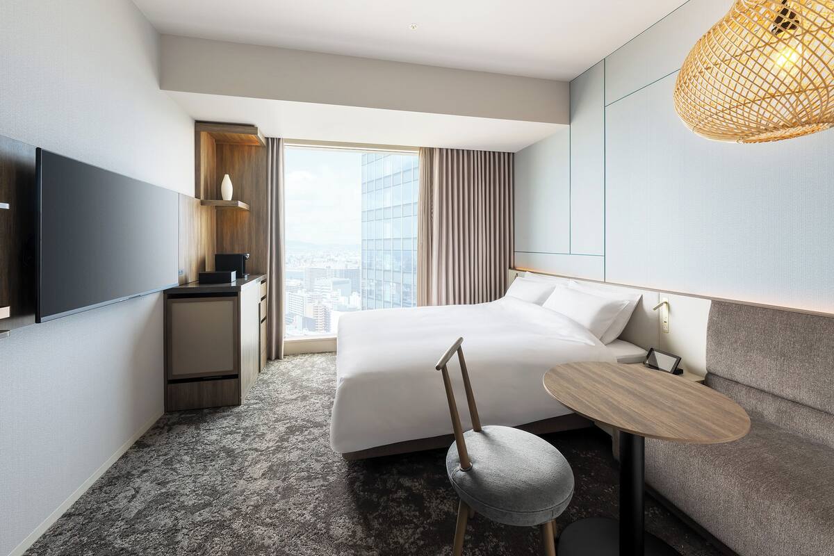 Quarto de hotel moderno com uma cama grande, uma pequena escrivaninha, uma cadeira, televisão e uma janela ampla com vista para a cidade.