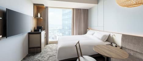 A modern hotel room with a large bed, a small desk, a chair, a television, and a large window with a city view.
