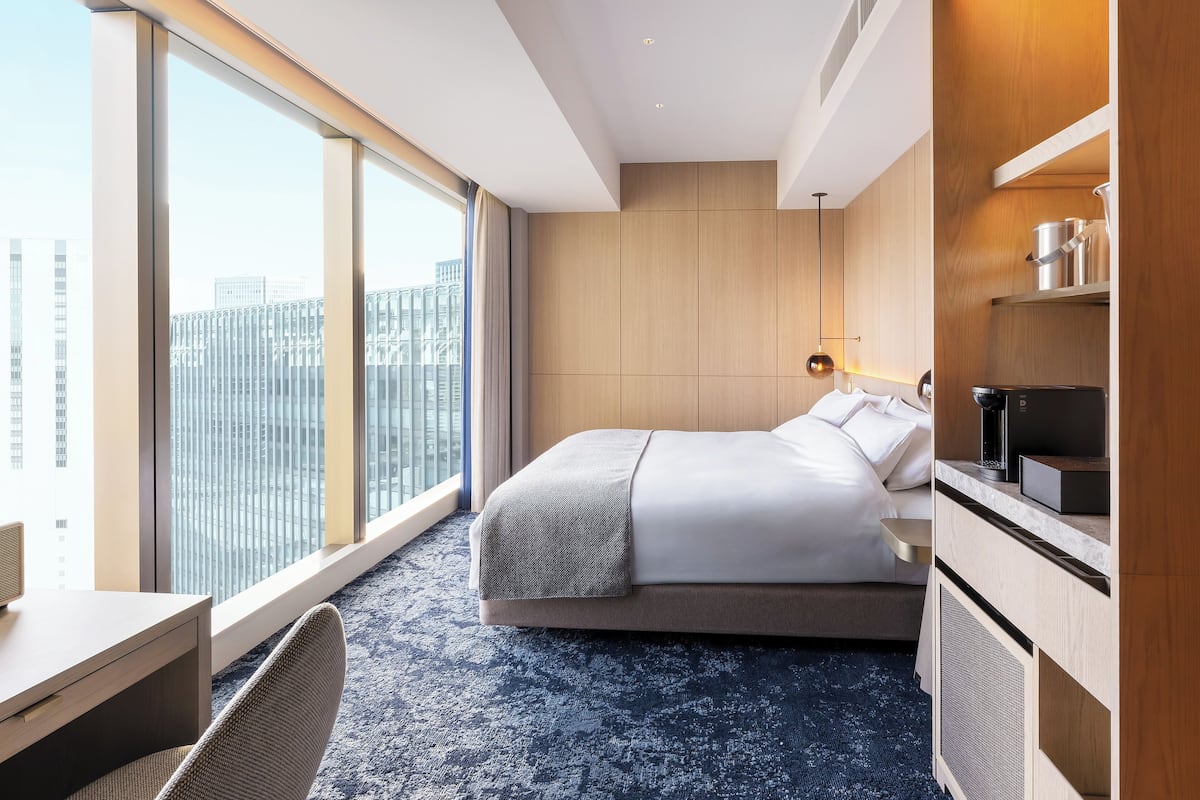Quarto de hotel moderno com uma janela grande, cama, escrivaninha e cozinha planejada.