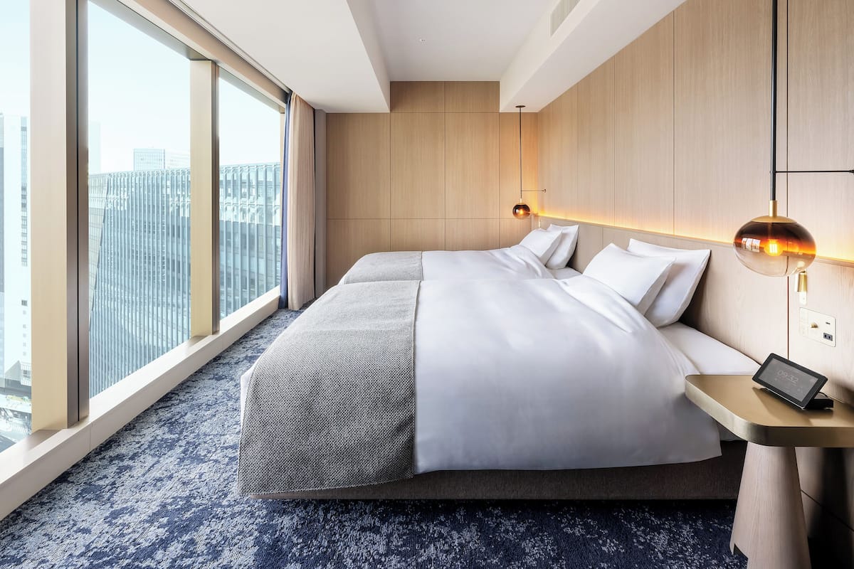 Quarto de hotel moderno com uma cama grande, uma mesinha de cabeceira com abajur e vista para a cidade através de amplas janelas.