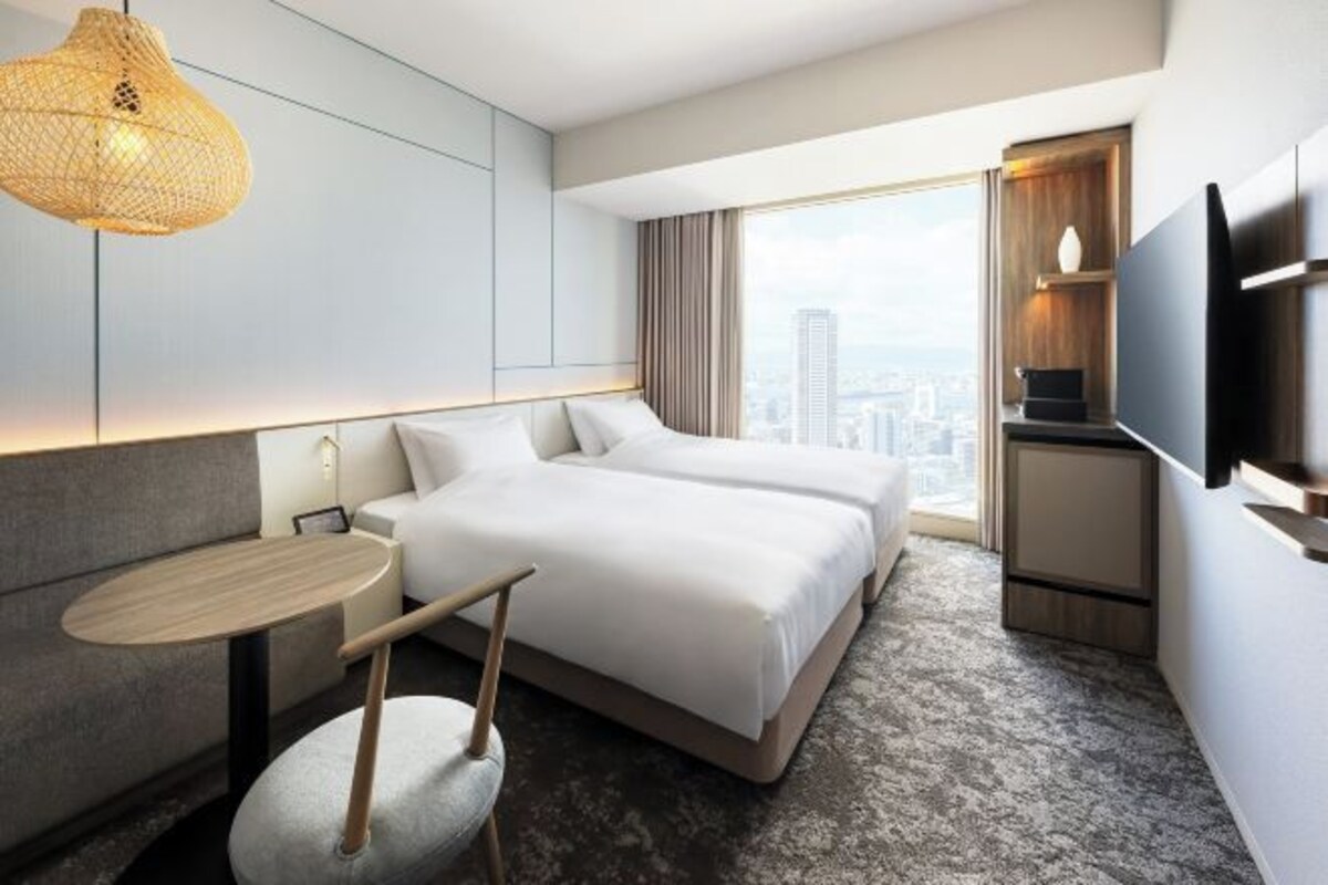 A modern hotel room with a large bed, a small table, a chair, a TV, and a city view.