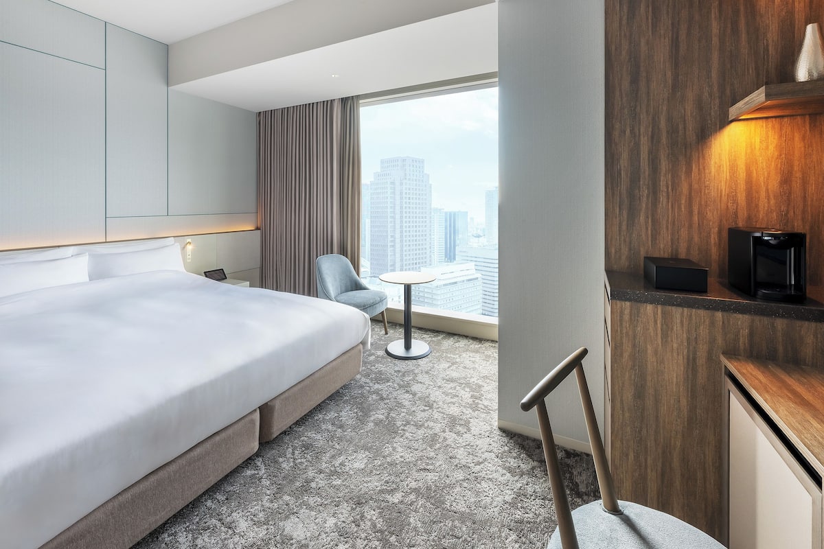 Quarto de hotel moderno com uma cama grande, uma mesa redonda pequena, uma cadeira e vista para a cidade através de uma janela ampla.