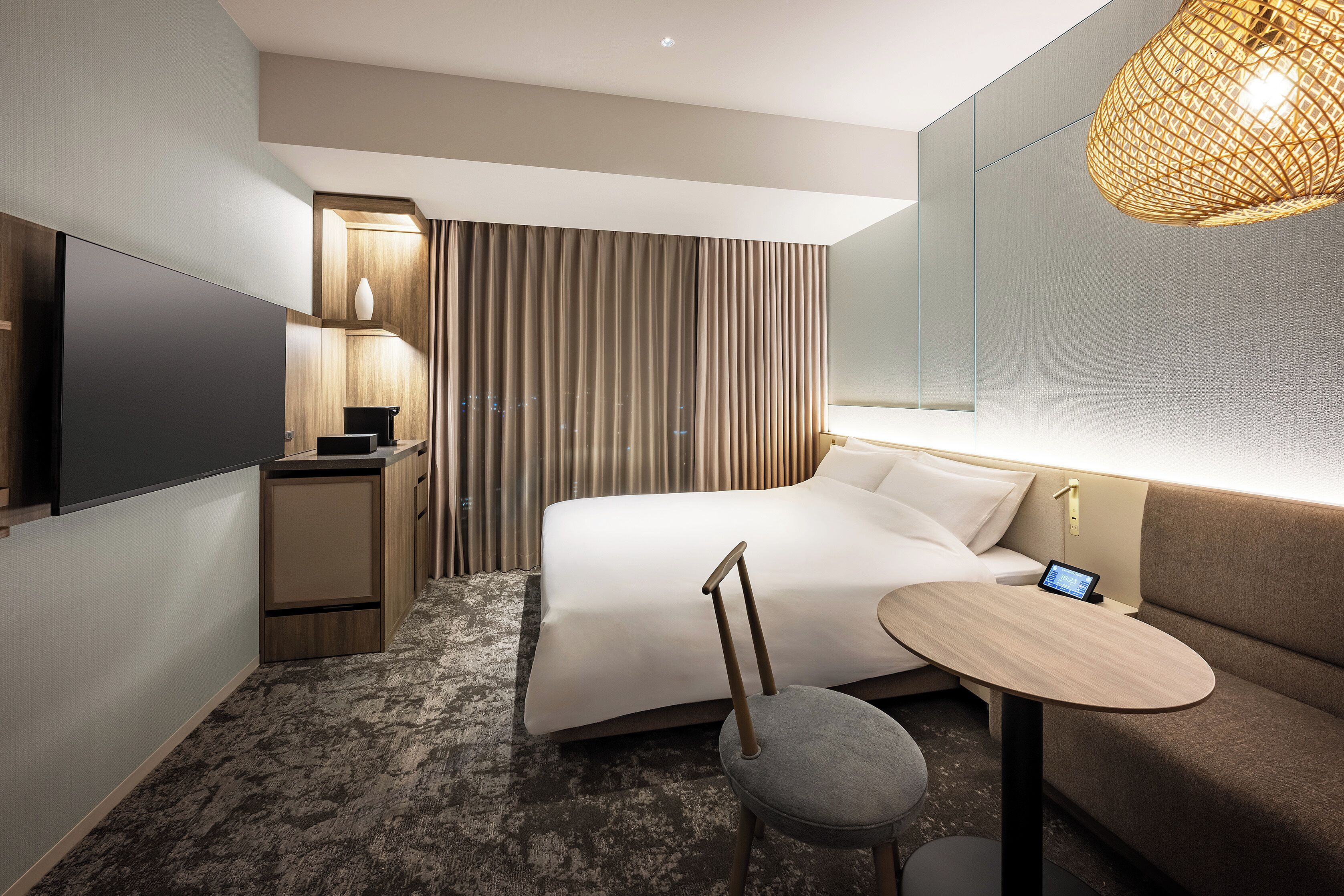 A modern hotel room with a bed, bedside table, TV, and a small seating area with a table and chair.