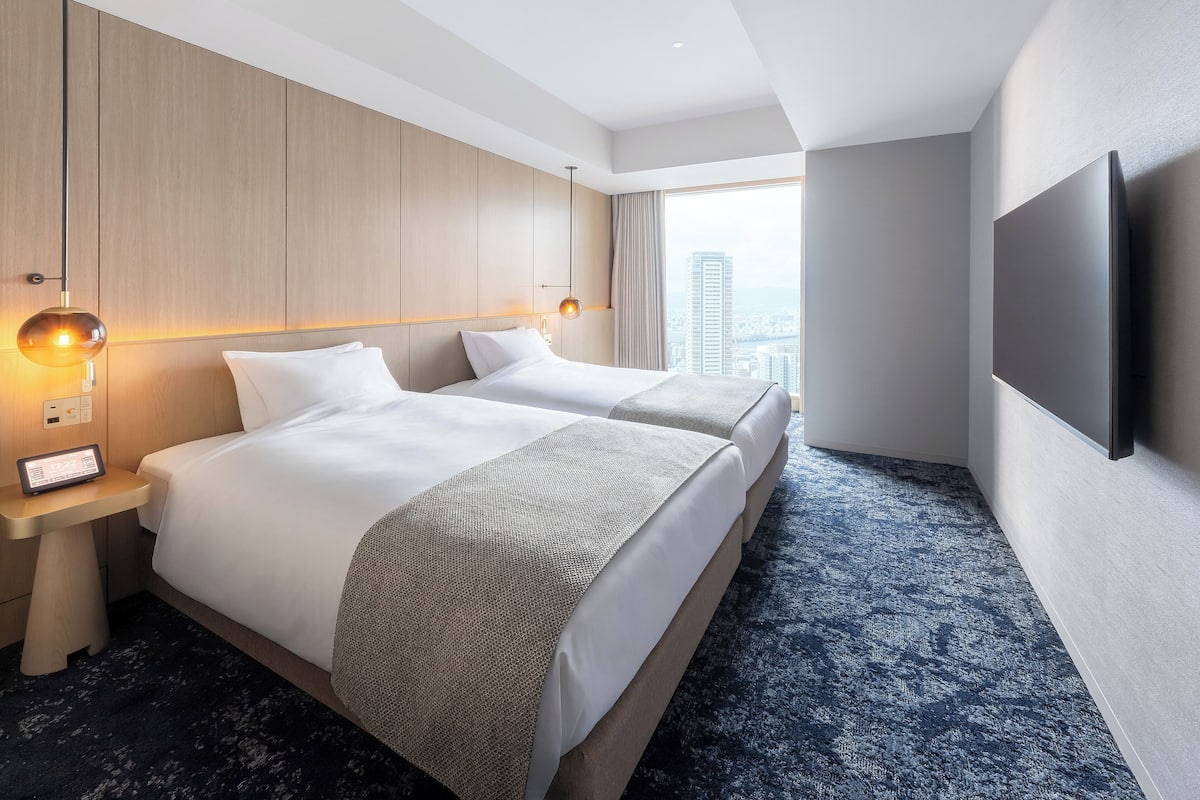Quarto de hotel com duas camas, uma TV de tela plana, mesa de cabeceira com abajur e vista para a cidade.