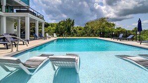 En klarblå pool med solstolar och parasoller, omgiven av en vit byggnad och träd.
