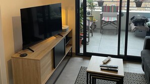 Ein Wohnzimmer mit einem Flachbildfernseher auf einem Holz-TV-Schrank, einem Couchtisch mit Fernbedienungen und einer Glastür, die auf einen Balkon mit Gartenmöbeln und Pflanzen führt.