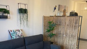 Ein modernes Wohnzimmer mit einem dunkelgrauen Sofa, einem runden Couchtisch aus Holz und einem gemusterten Teppich. Zwei Zimmerpflanzen sind zu sehen, eine auf dem Sofa und die andere hinter einem Bambusbildschirm.