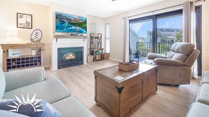 A spacious living room with a fireplace, a large flat-screen TV, a ceiling fan, and a balcony with a view of palm trees. - Exclusive End-Unit! Sea Winds #36: Gated Ocean Views & Direct Beach Access! (St. Augustine Beach)
