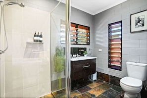 A modern bathroom with a glass shower enclosure, a dark wood vanity, a white toilet, and a unique window with multiple colored panels. - Relax at Sudbury Shores Beach House- Tropical Kewarra Beach Retreat | Lagoon Pool + Outdoor Bath (Kewarra Beach)