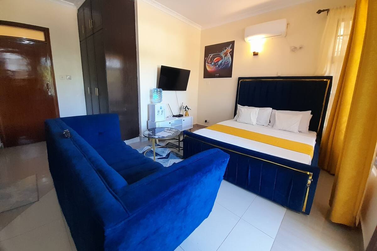 Quarto de hotel com cama, poltrona azul, pequena mesa com dispensador de água e televisão fixada na parede.