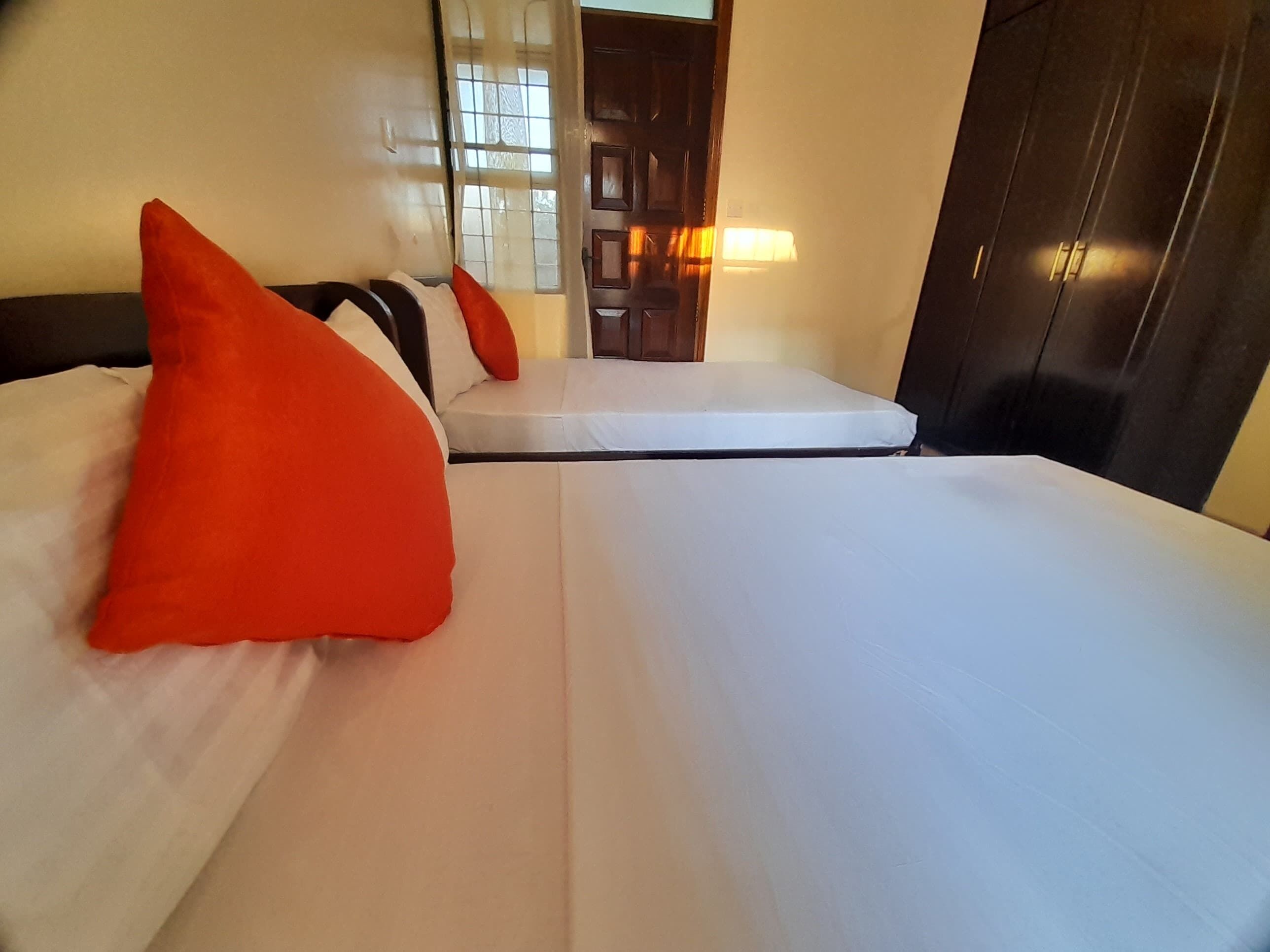 Habitación de hotel con dos camas, cada una con ropa de cama blanca y almohadas rojas. Hay una puerta de madera y un espejo en la pared.