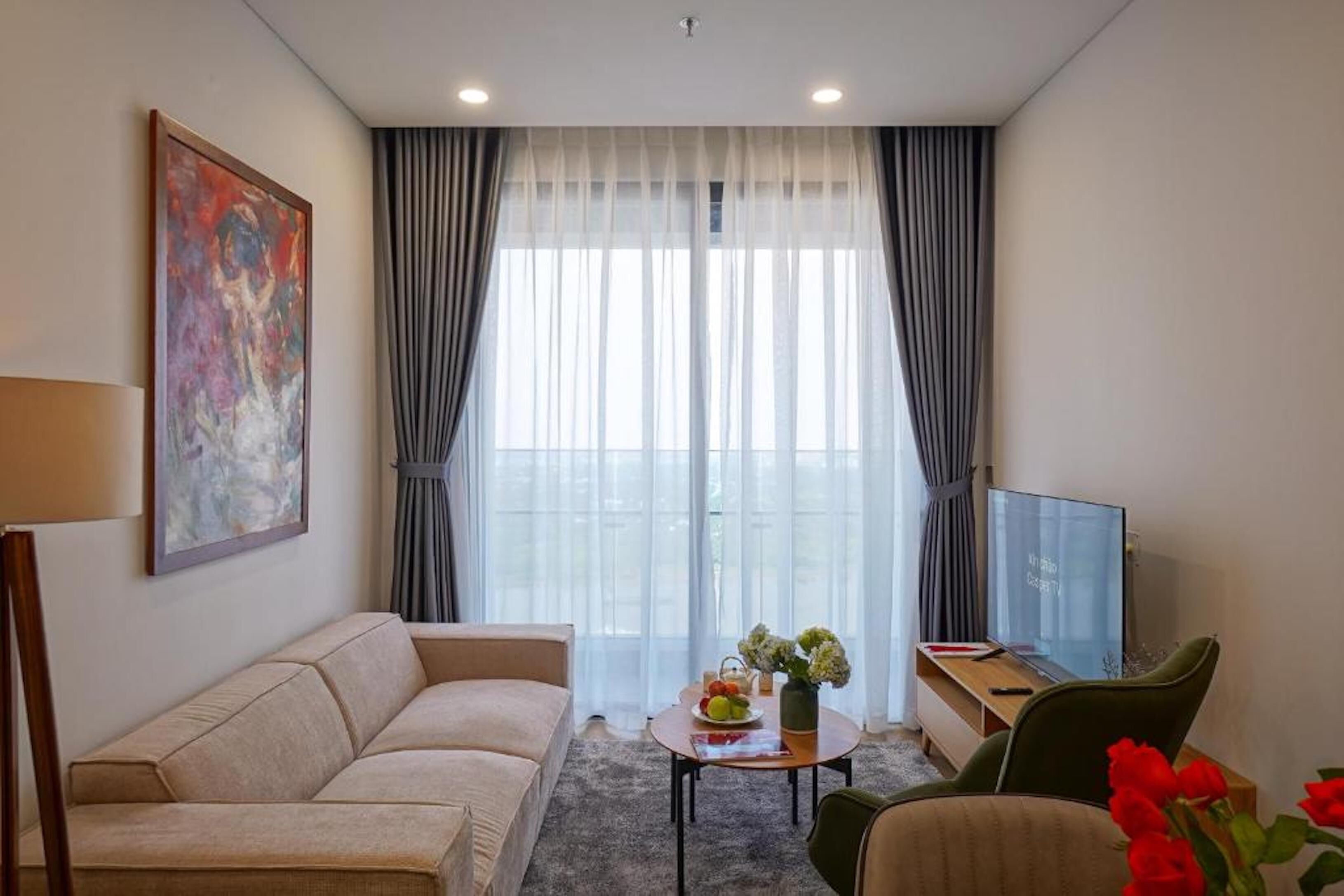 A modern hotel room with a sofa, a small table with a vase and flowers, a desk with a computer, and a large window with curtains.