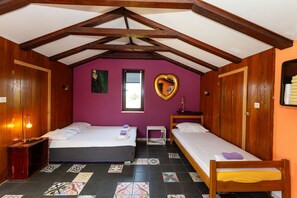 A room with two beds, wooden beams, a purple wall, a heart-shaped mirror, and a window.