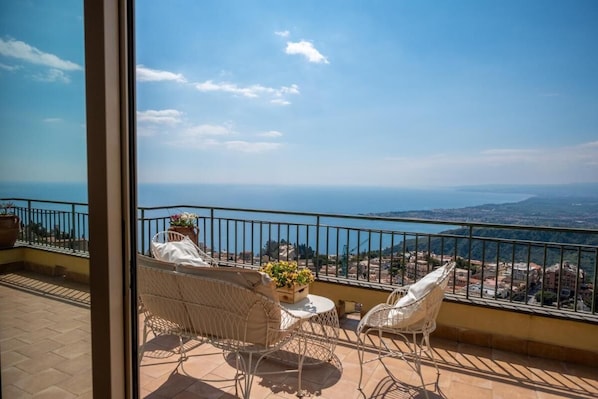 A balcony with wicker chairs and a small table, overlooking a coastal cityscape.