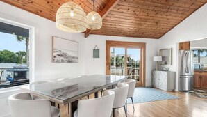 A modern kitchen with a large island, white chairs, a wooden ceiling, and a view through a glass door.