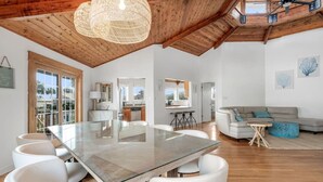 A spacious living area with a glass-top dining table, white chairs, a grey sofa, and a woven ceiling light fixture.