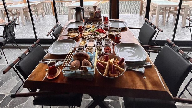 A dining table set with various dishes and drinks, surrounded by chairs, with a view of mountains through large windows. - Pagen Bungalov (Hopa)