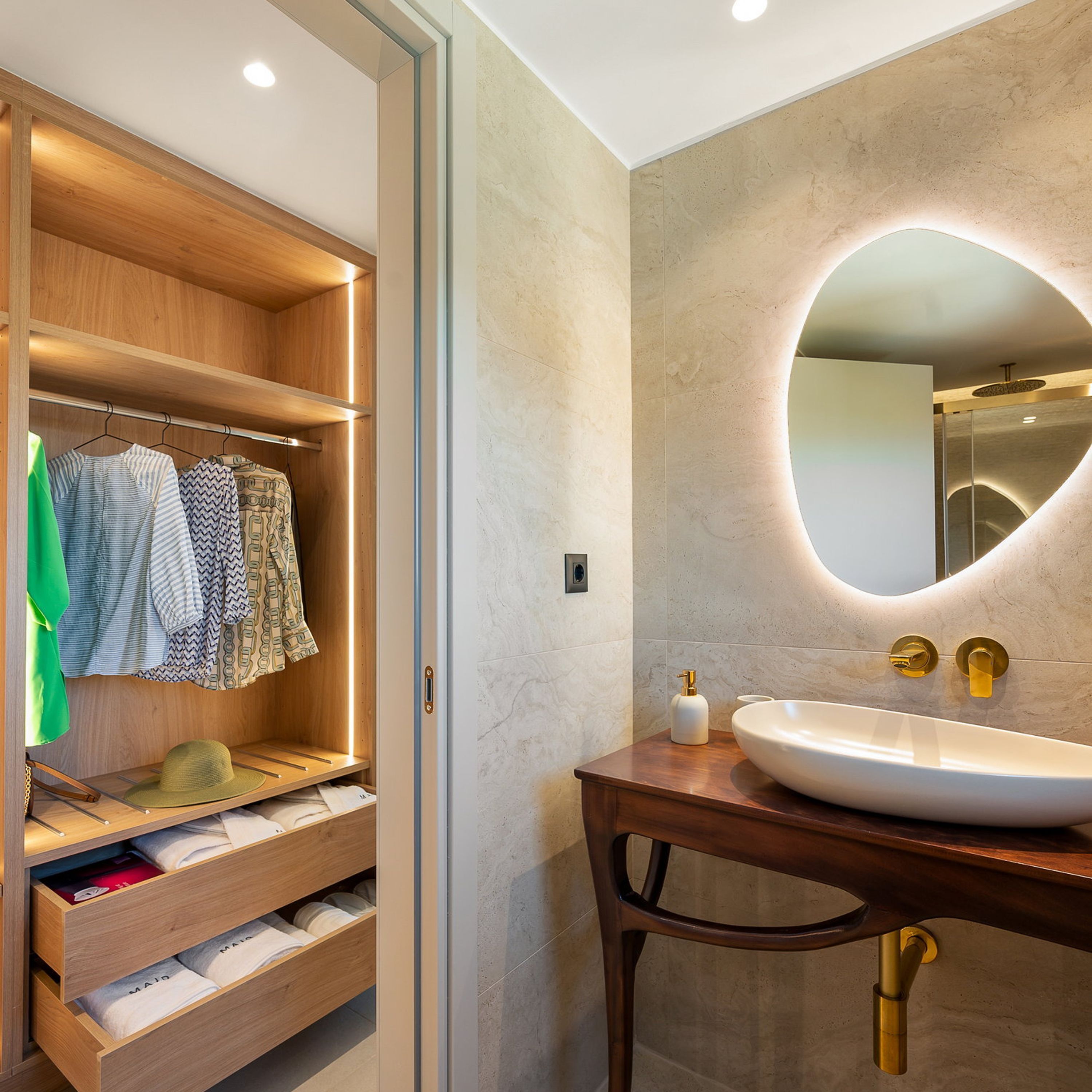 A modern bathroom with a wooden vanity, a large oval mirror, and a built-in wardrobe with hanging clothes and shelves.