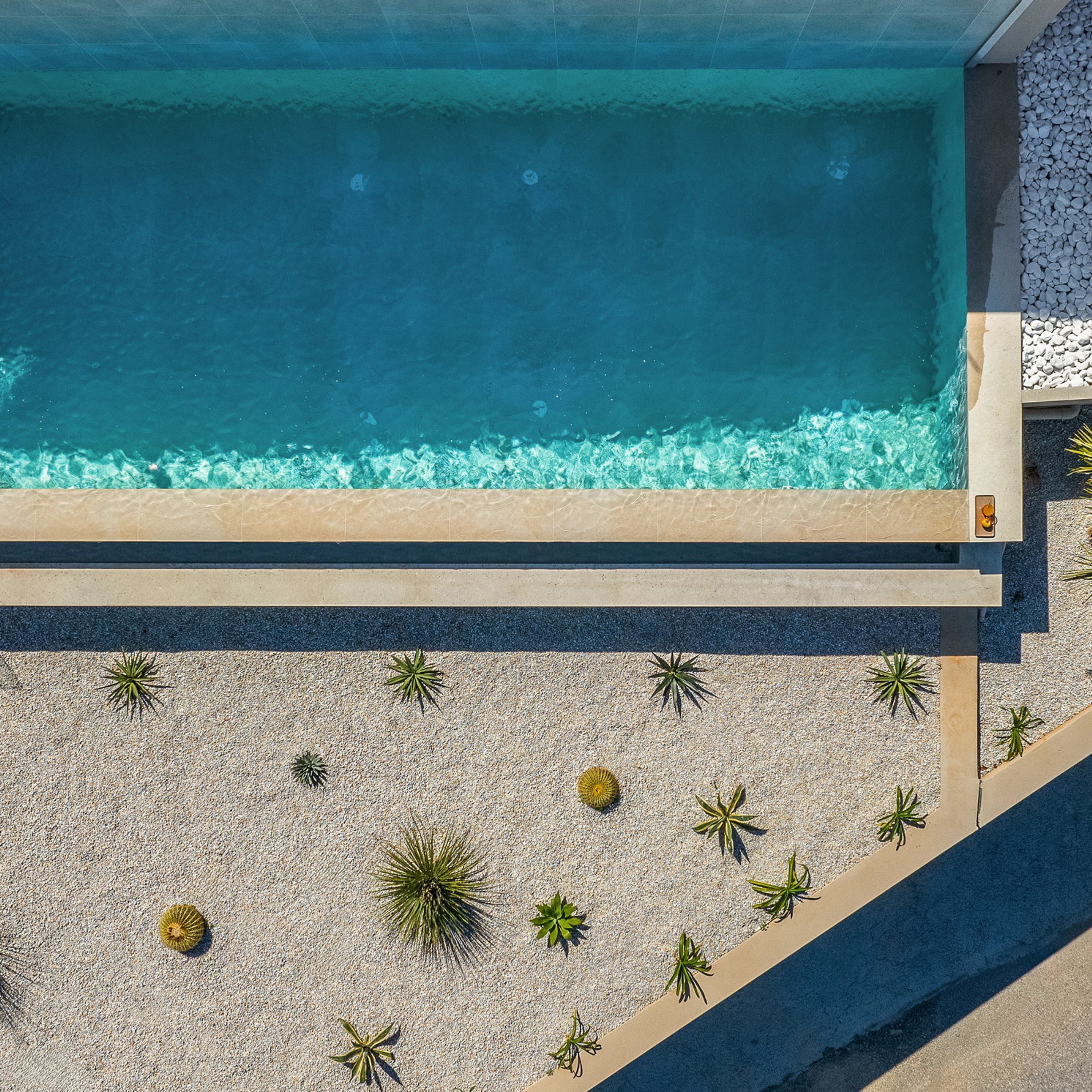 An aerial view of a swimming pool with clear blue water, surrounded by a pebble beach and small green plants.