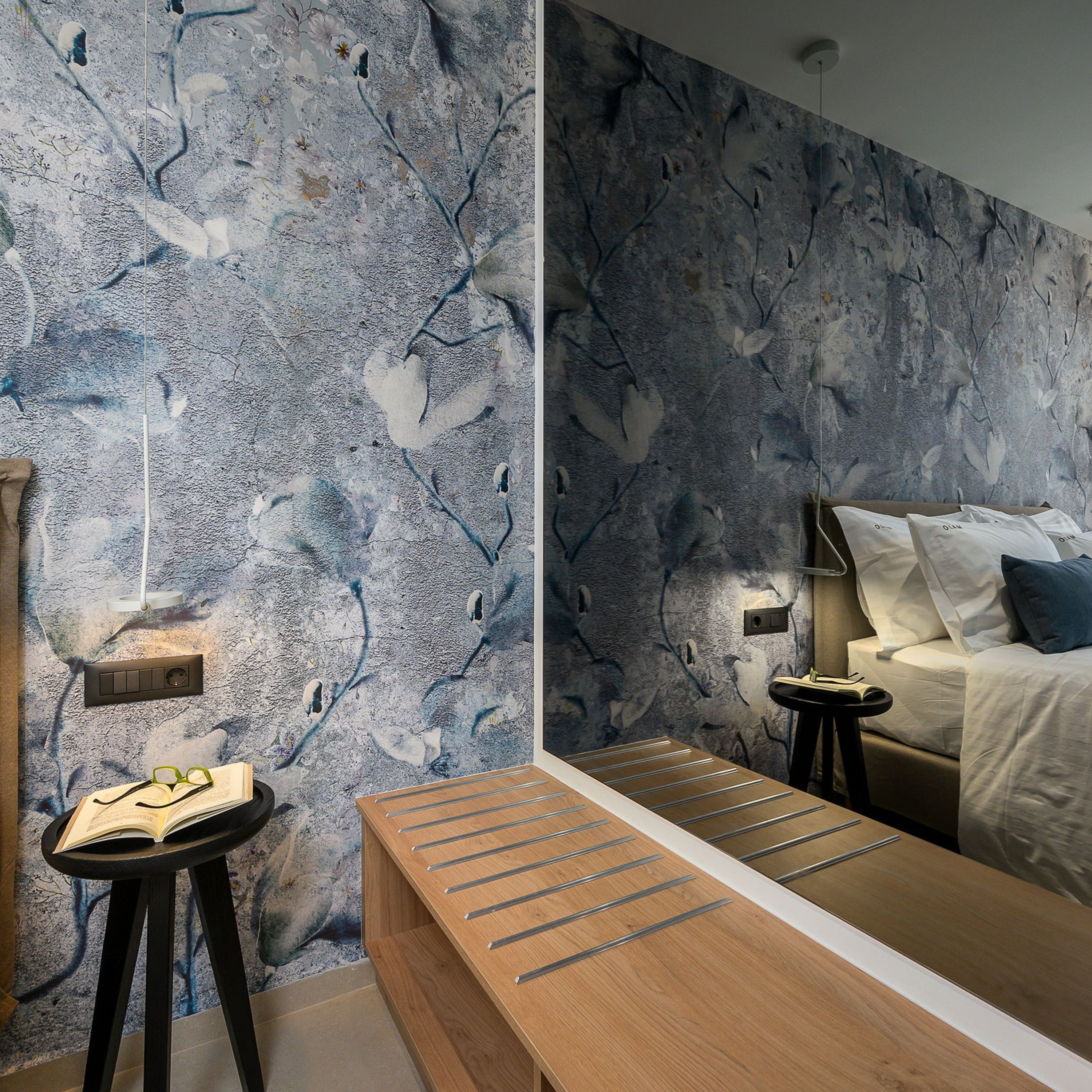 Ein modernes Schlafzimmer mit strukturierter Wand, einem Nachttisch aus Holz, einem Bett mit weißer Bettwäsche und einem kleinen Nachttisch mit Lampe.
