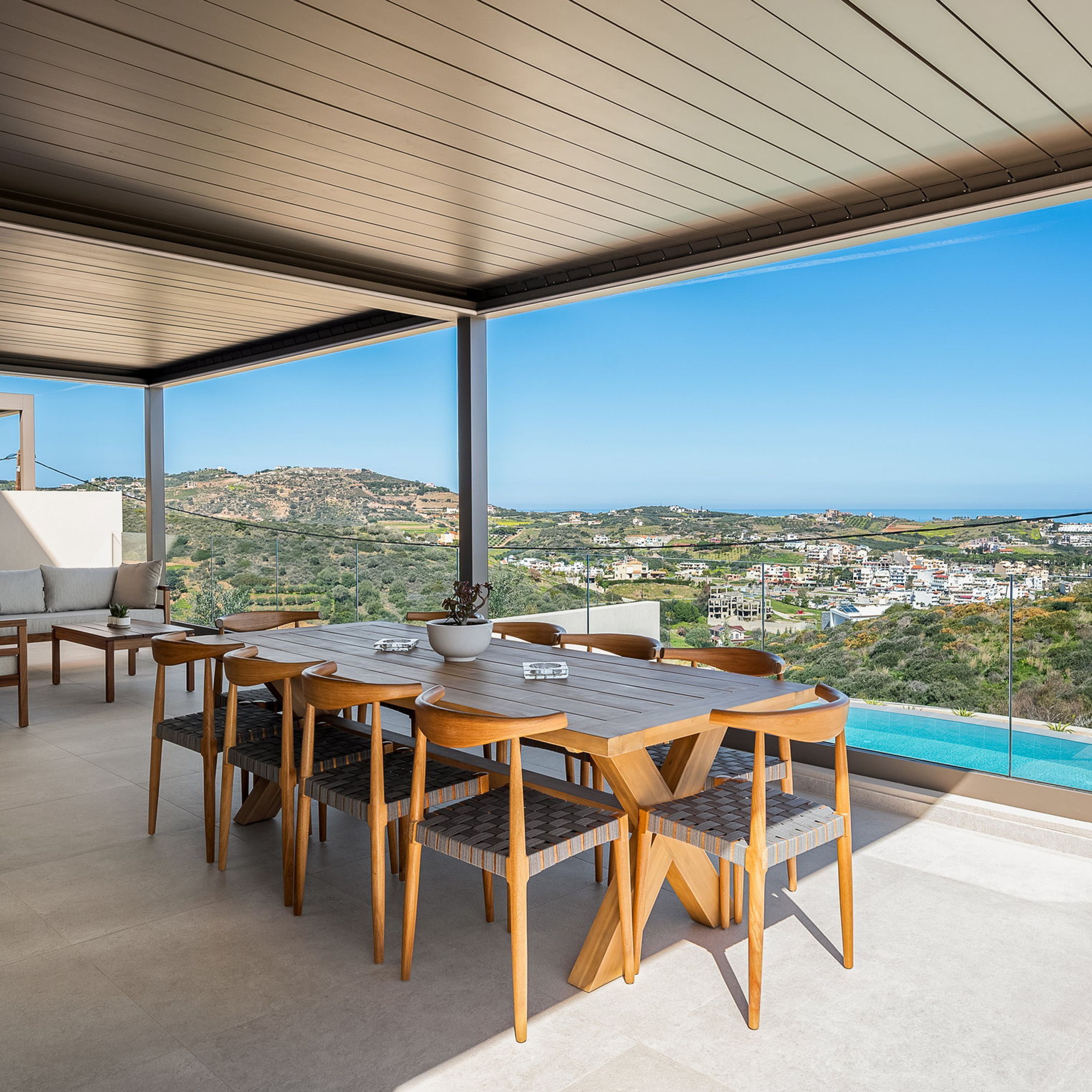 A terrace with a wooden table and chairs, a potted plant, and a view of a coastal town.