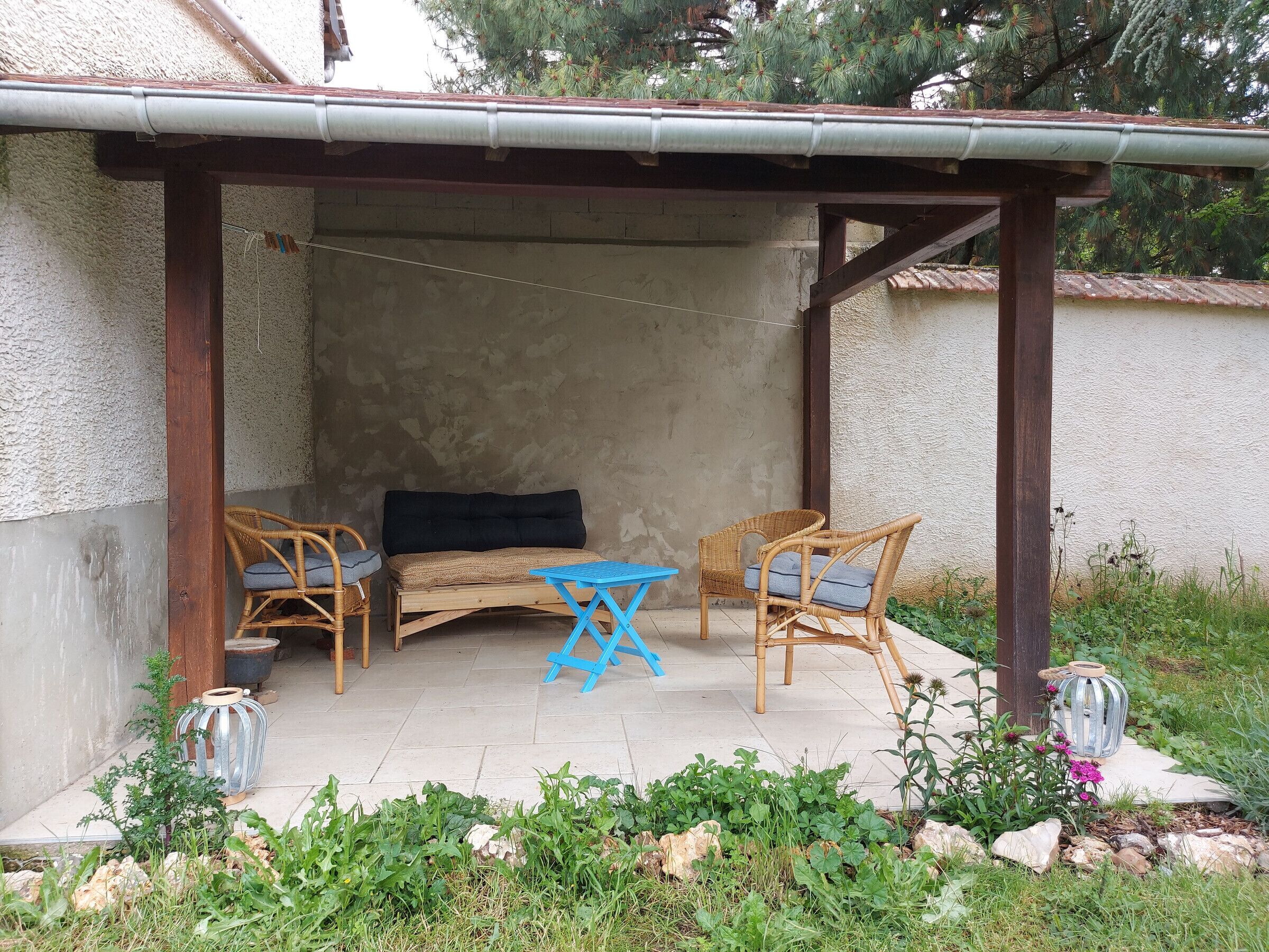 Un espace extérieur aménagé, abrité, avec un canapé et deux fauteuils en osier, une petite table bleue et une lanterne en métal.