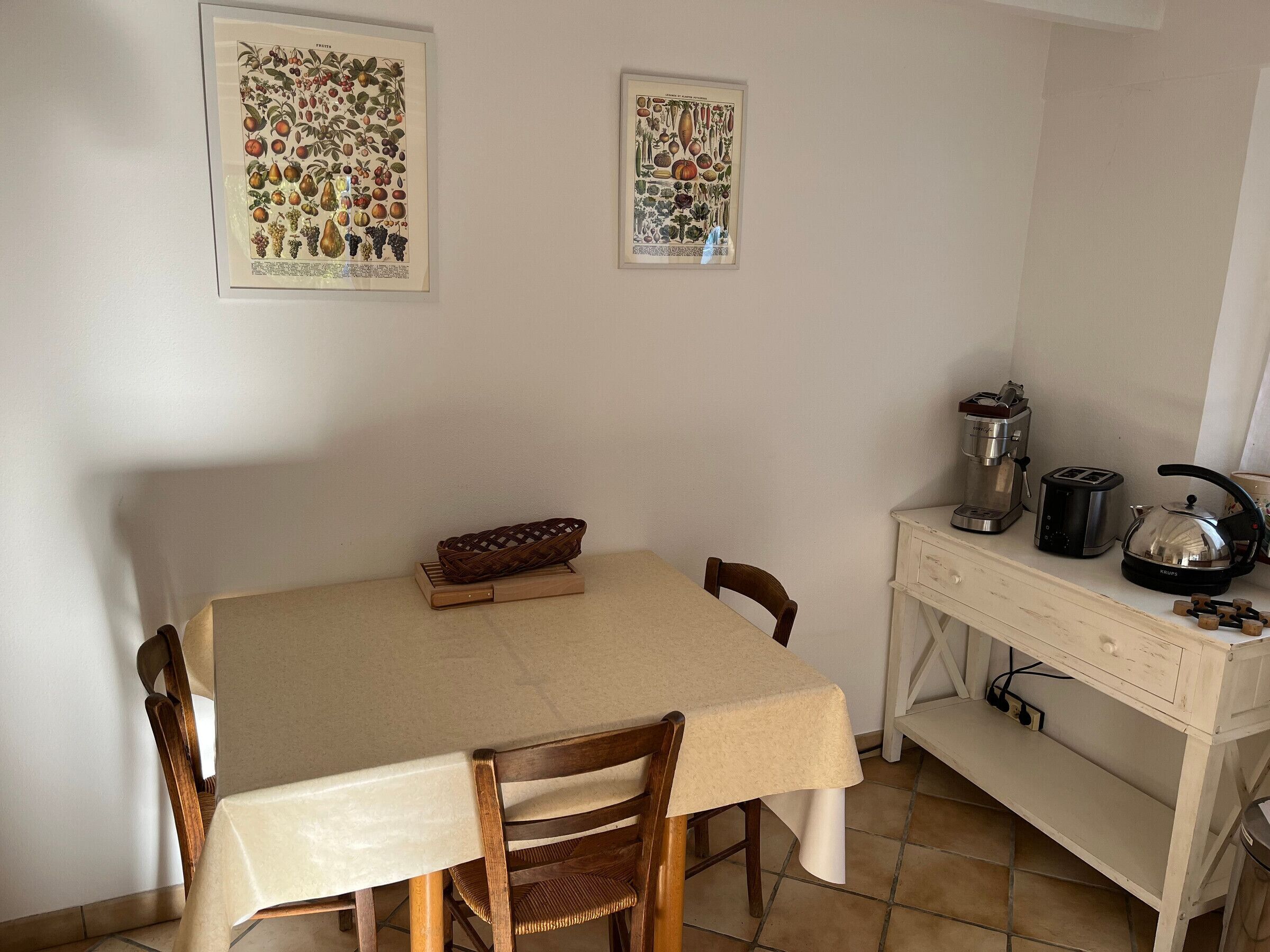 Ein Essbereich mit Tisch, Stühlen, einer Kaffeemaschine, einem Wasserkocher und zwei gerahmten Pflanzenbildern an der Wand.
