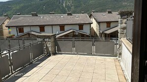 Une cour dallée, un mur en pierre et des bâtiments avec des toits en tuiles et des volets en bois.