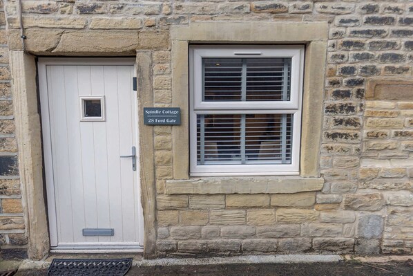 A stone building with a white door and a window with shutters. A sign above the door reads "Spindle Cottage 28 Ford Gate."