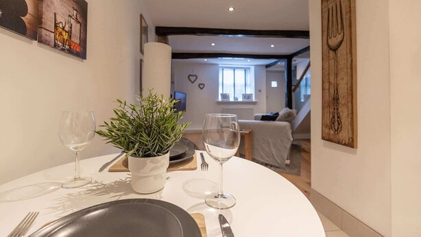 Een eetgedeelte met een ronde tafel voor twee personen, een potplant en een uitzicht op een woonkamer met een bank en een trap.