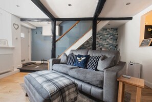 Uma sala de estar moderna com um sofá cinza, um banquinho xadrez e uma mesa lateral de madeira. Uma escada com parede azul e corrimão de madeira é visível ao fundo.