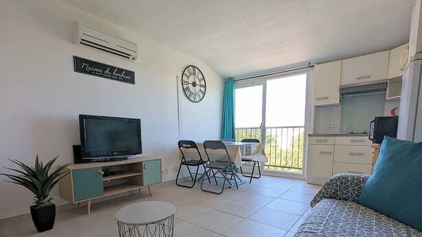 Un espace de vie compact comprenant un téléviseur, une table à manger, des chaises, une petite cuisine et un balcon avec des rideaux.