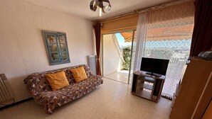 Ein Wohnzimmer mit einem gemusterten Sofa, einem Fernseher auf einem Holzboden und Blick durch eine Glastür.