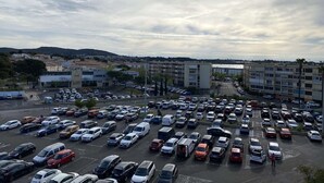 En stor parkeringsplads med flere rækker parkerede biler, omgivet af bygninger og et bakket landskab i baggrunden.