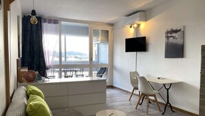 En moderne stue med sofa, spisebord, stole, fjernsyn og et stort vindue med gardiner.