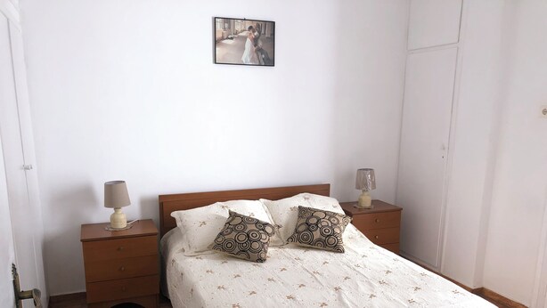 A bedroom with a wooden bed, white bedding, two patterned pillows, a nightstand with a lamp, and a small table with another lamp.