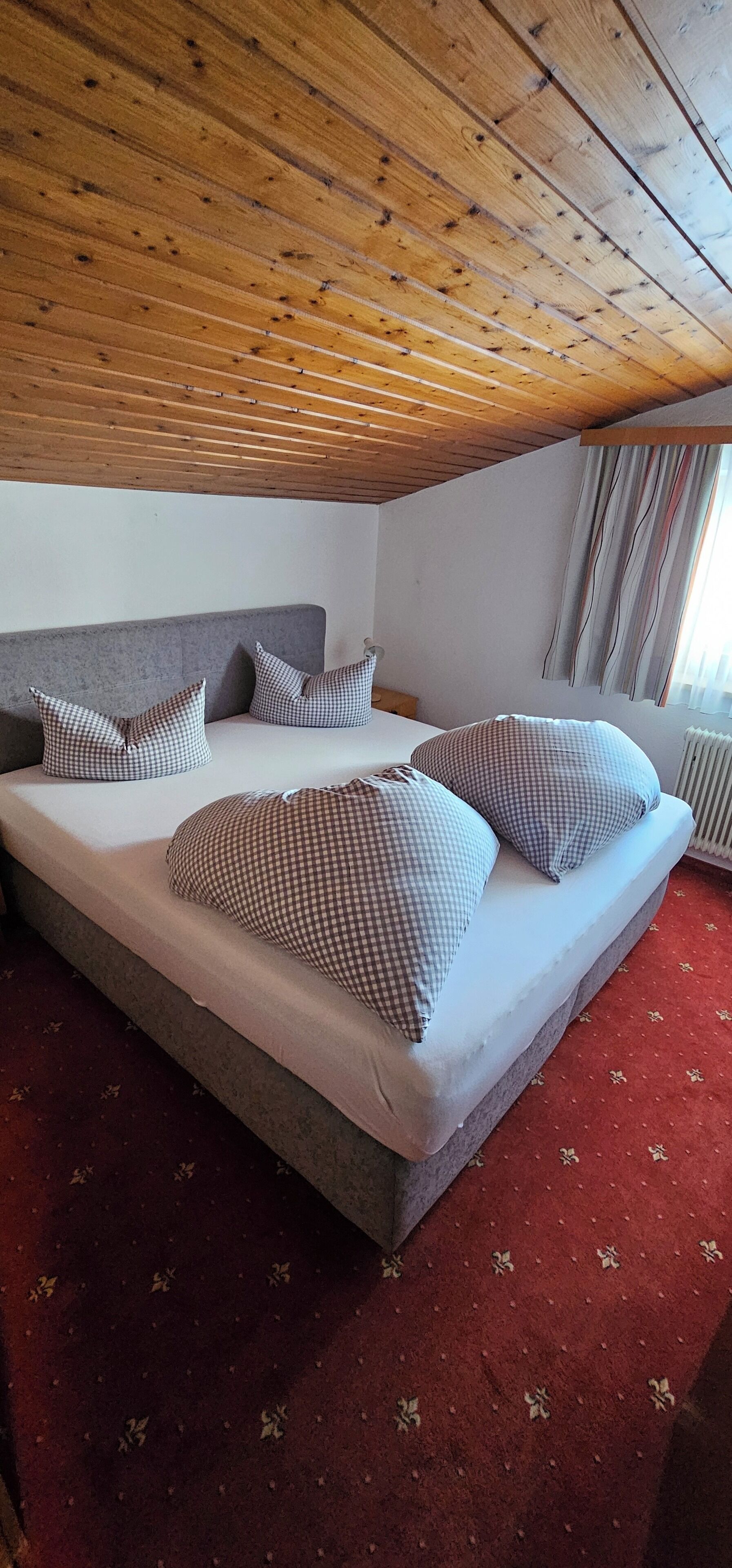 A bedroom with a wooden ceiling, a grey headboard, a bed with checkered bedding, and a red carpet.