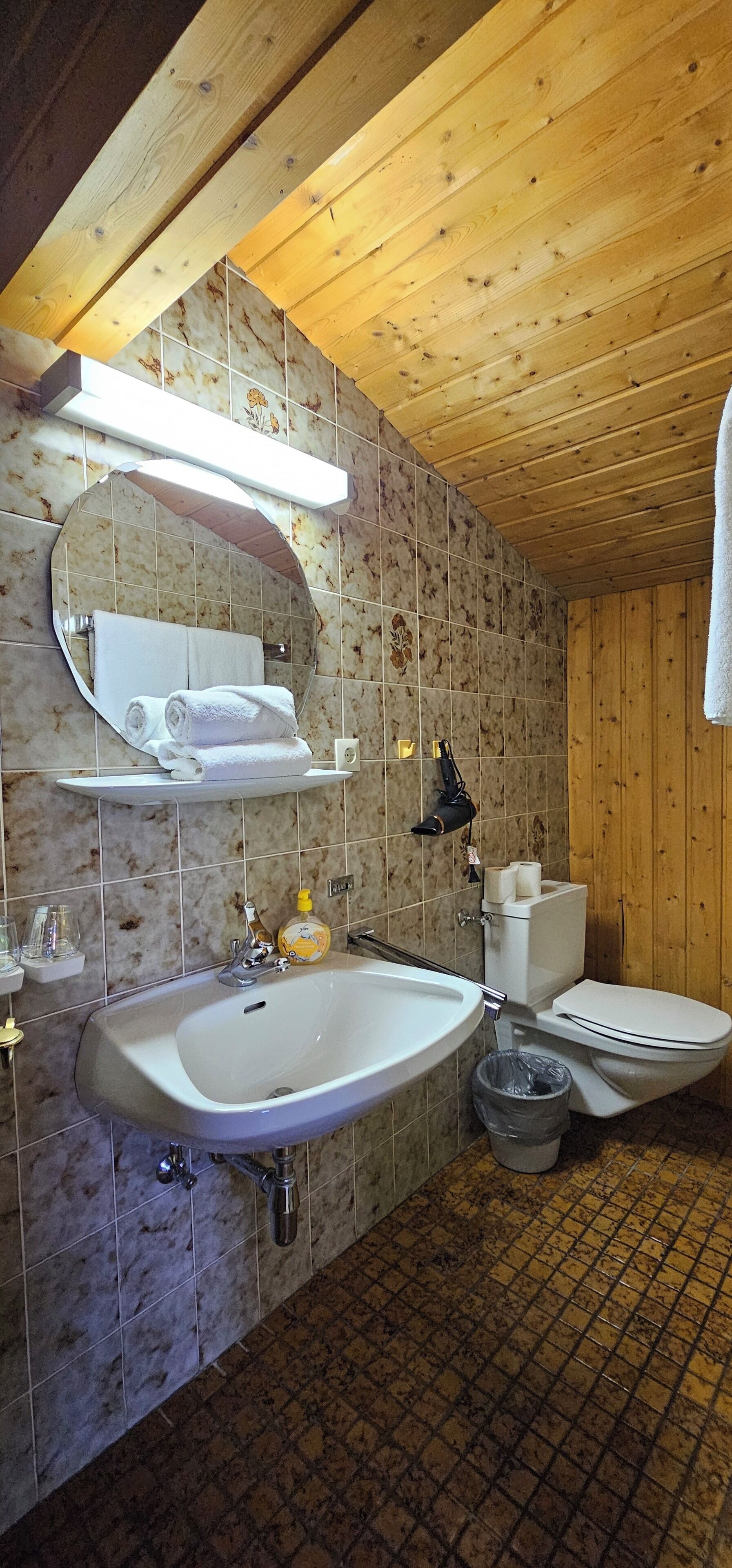 A bathroom with a wooden ceiling, a large mirror, a sink, a toilet, and a trash bin.
