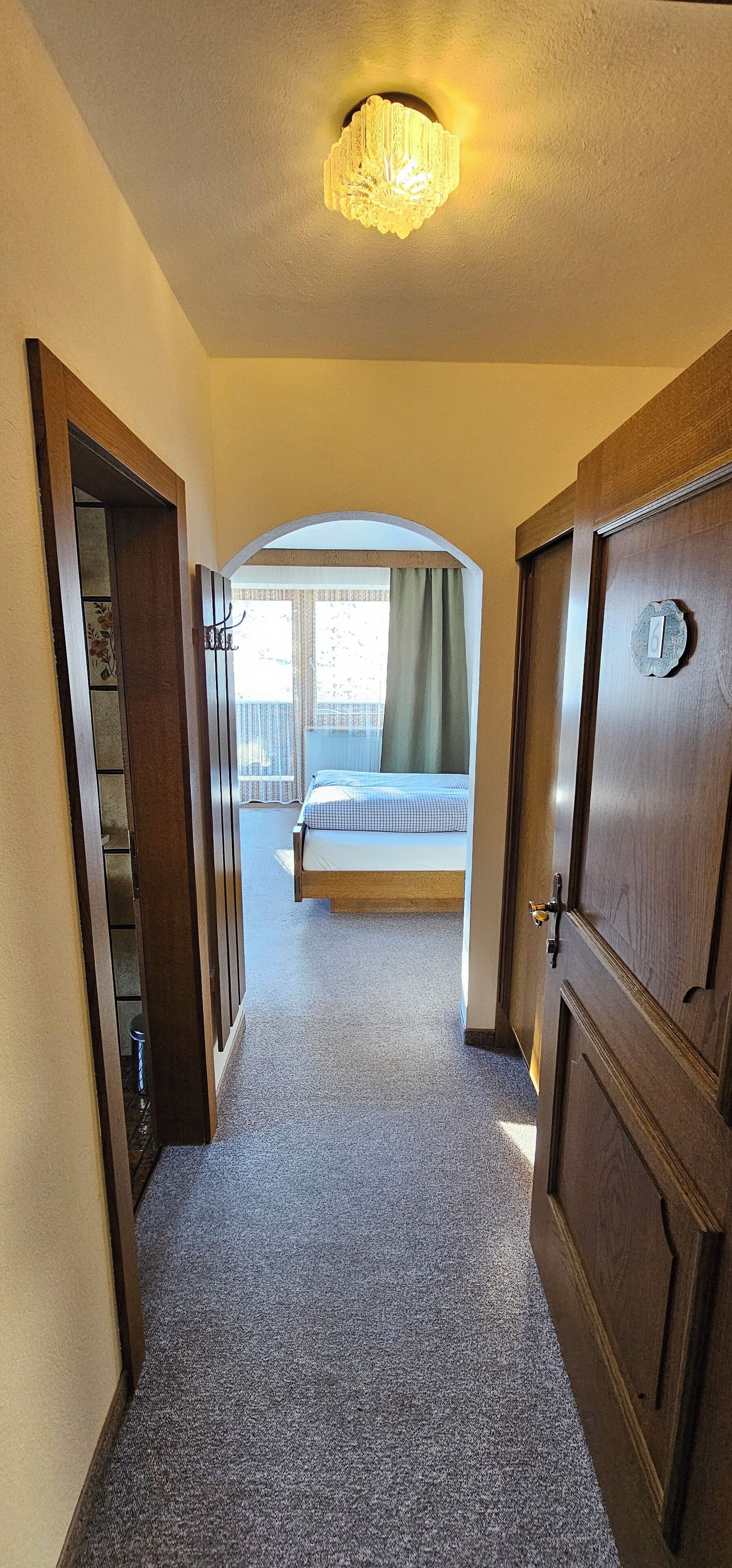 Pasillo de hotel que lleva a un dormitorio con cama, ventana con cortinas y un candelabro.