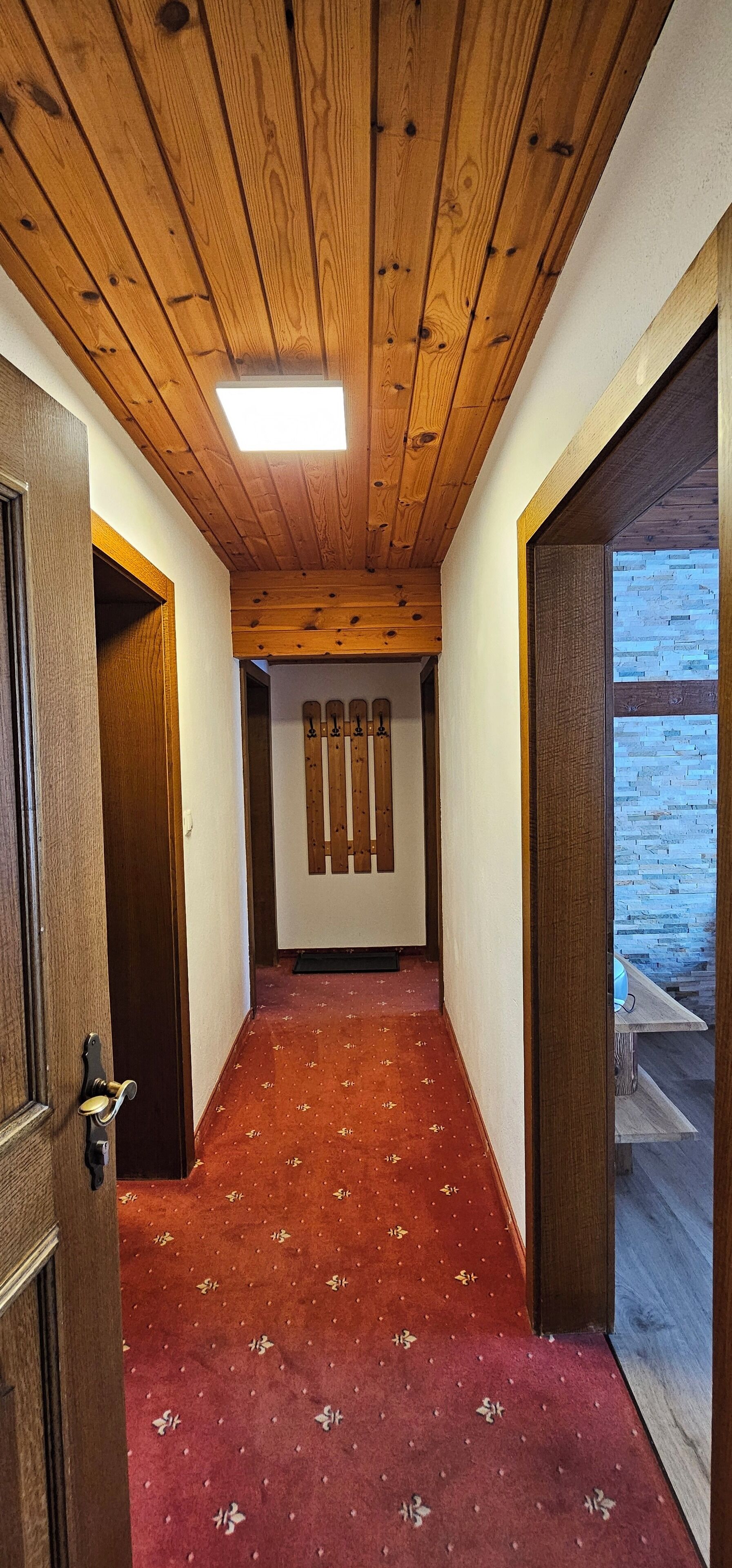 Un estrecho pasillo con vigas de techo de madera, una alfombra roja con motivos florales y vistas a una terraza de madera azul a través de un puerta de cristal.