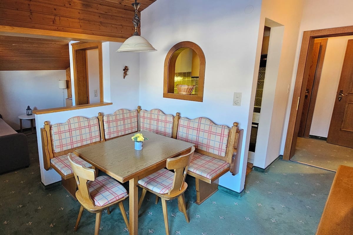 A dining area with a wooden table and plaid upholstered chairs, a small side table with a lamp, and a mirror above the table.