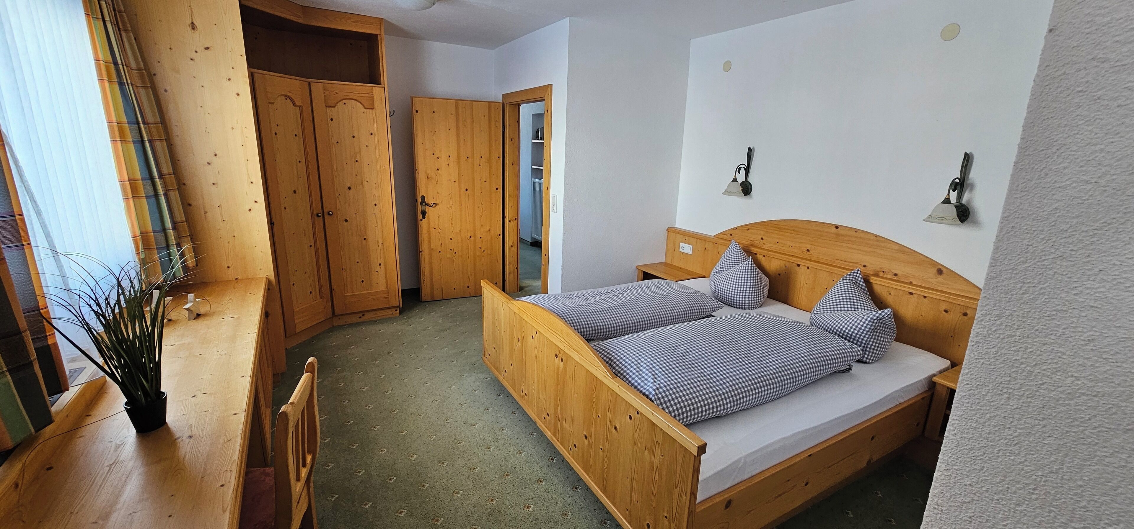 A bedroom with a wooden bed, checkered pillows, a nightstand, a wooden wardrobe, and a potted plant.